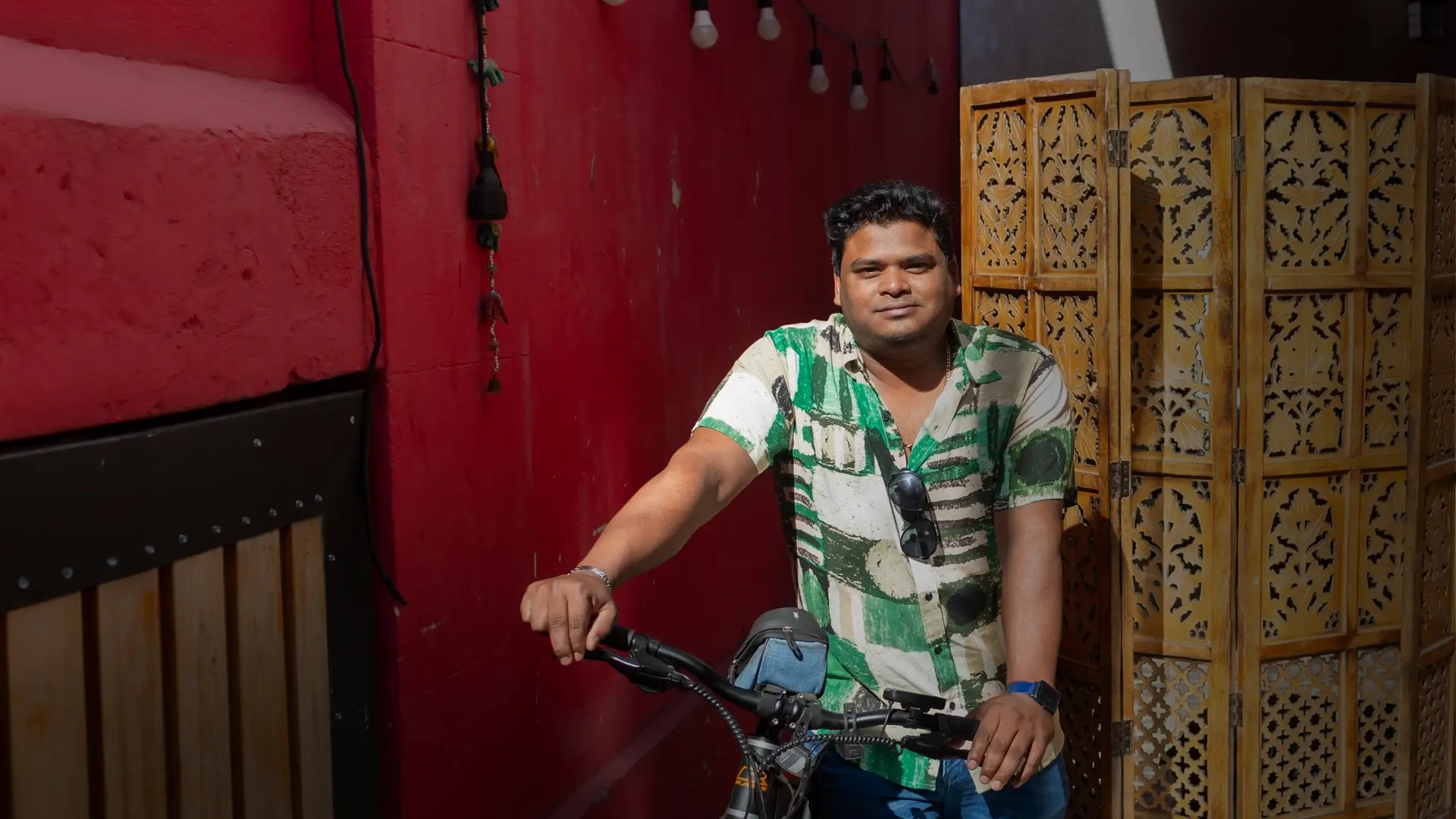 The image features a man standing next to a parked motorcycle. He is wearing a patterned shirt and appears to be posing for a picture. The motorcycle is positioned in front of a red wall, which serves as a backdrop for the scene.