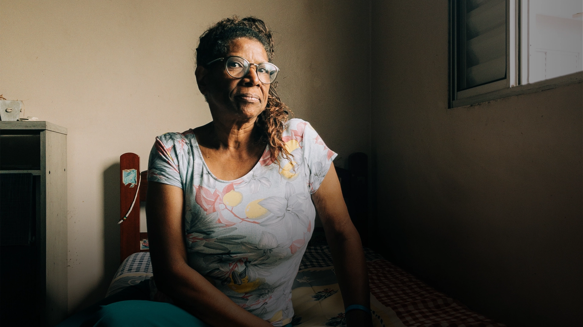 The image features a woman sitting on a bed in a room. She is wearing a floral print shirt and glasses. The room appears to be dimly lit, which adds to the overall atmosphere of the scene.