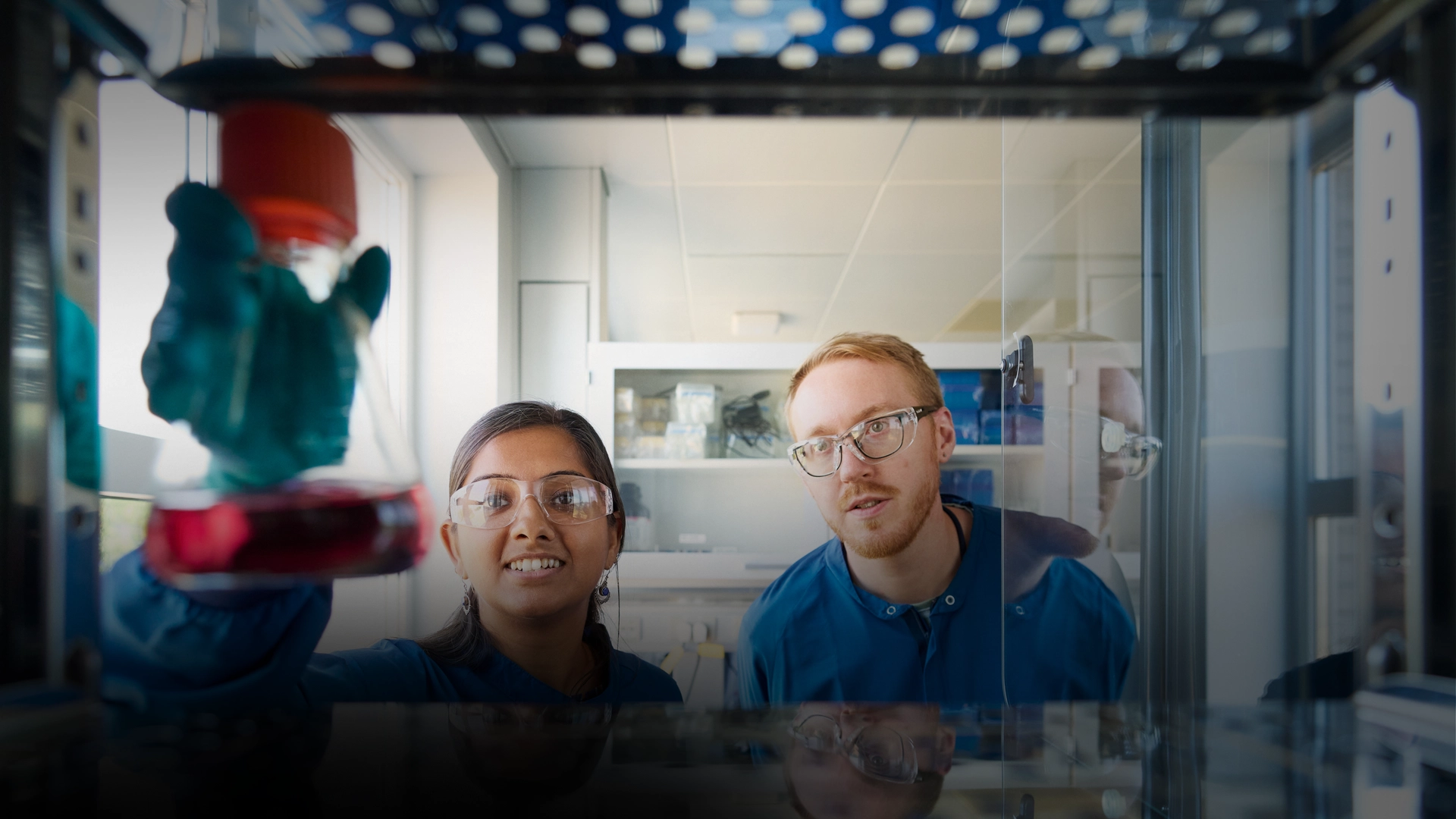 The image features a man and a woman in a lab setting, both wearing lab coats and goggles. They are standing in front of a blue counter, working together on an experiment or task. The woman is on the left side of the image, while the man is on the right side.
In the lab, there is a sink against the wall, and various items can be seen on the counter, such as a bottle on the left side and a cup on the right side. Additionally, there are two computer mice, one near the center of the counter and the other closer to the right edge. A pair of scissors is also present, located near the center of the counter.